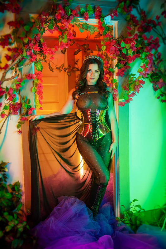 Model wearing a sheer black bodysuit with a holographic lace-up waist cincher and long flowing cape, standing in a doorway framed with pink flowers and colorful lighting.